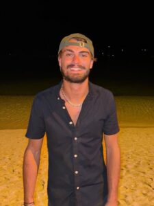 Un ciel étoilé, au bord de la mer, sur une plage de sable. Dan Elkayam, un jeune homme sourriant, porte une casquette verte et une chemise noire.