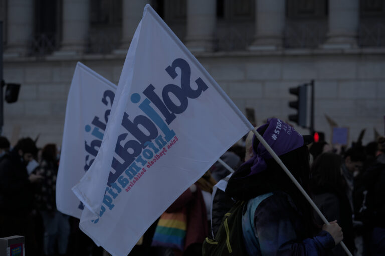 manifestation le jour avec des personnes qui marchent de gauche à groite on voit au premier plan une personne qui tient un drapeau solidaires informatique et au second plan un tote bag aux couleurs LGBTI